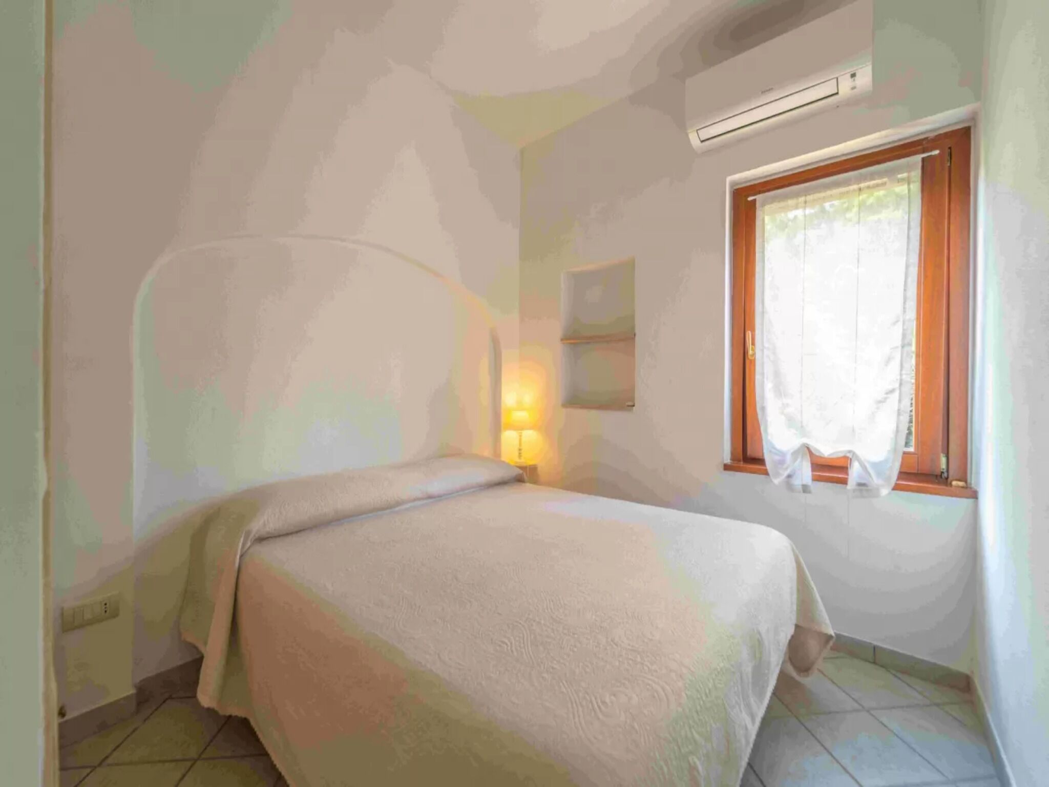Un dormitorio con cama, ventana con cortinas, aire acondicionado instalado en la pared y estante fijado a la pared.
