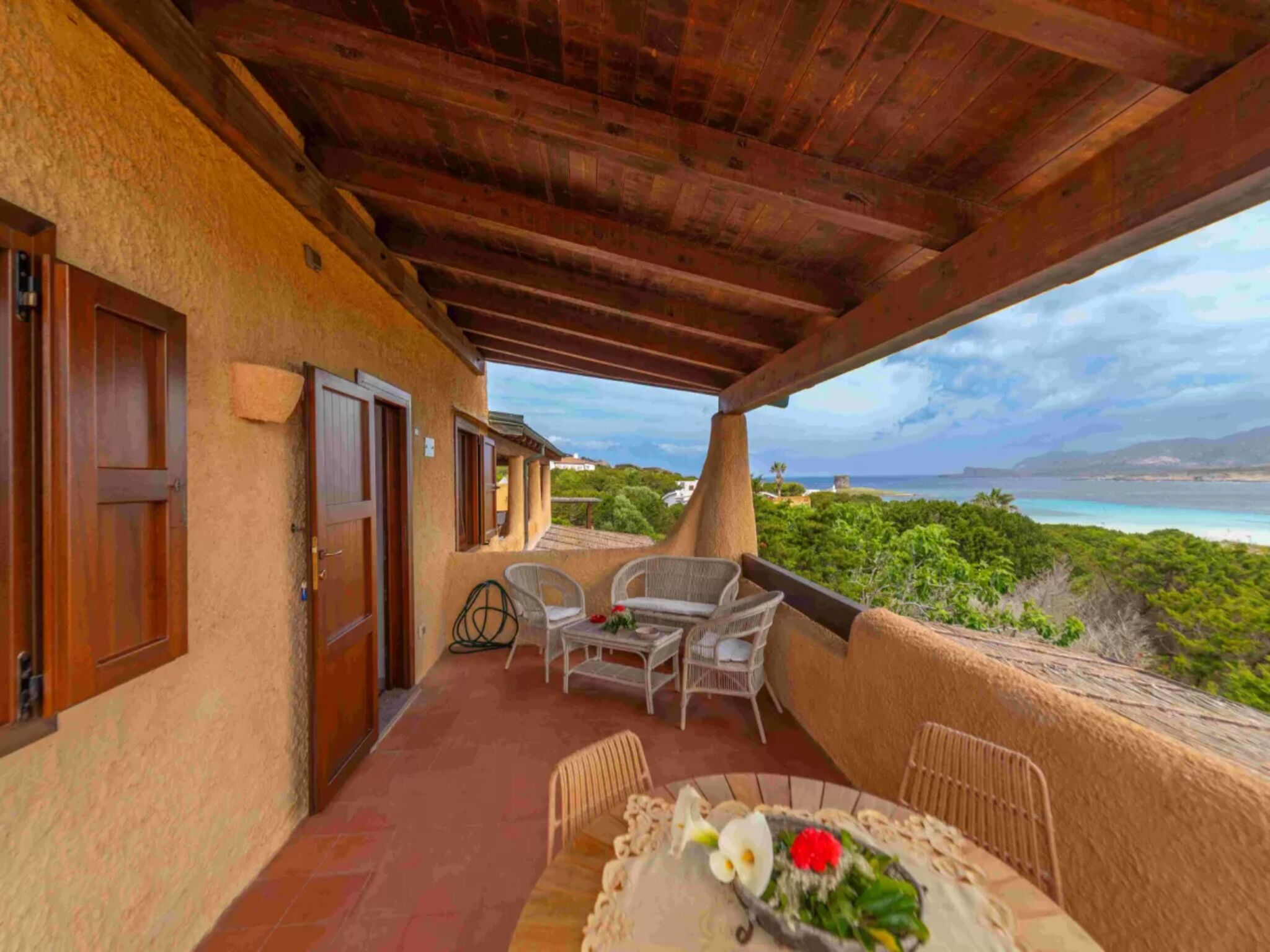 Uma área externa coberta com mesa e cadeiras, vista para o oceano e um edifício com uma porta.
