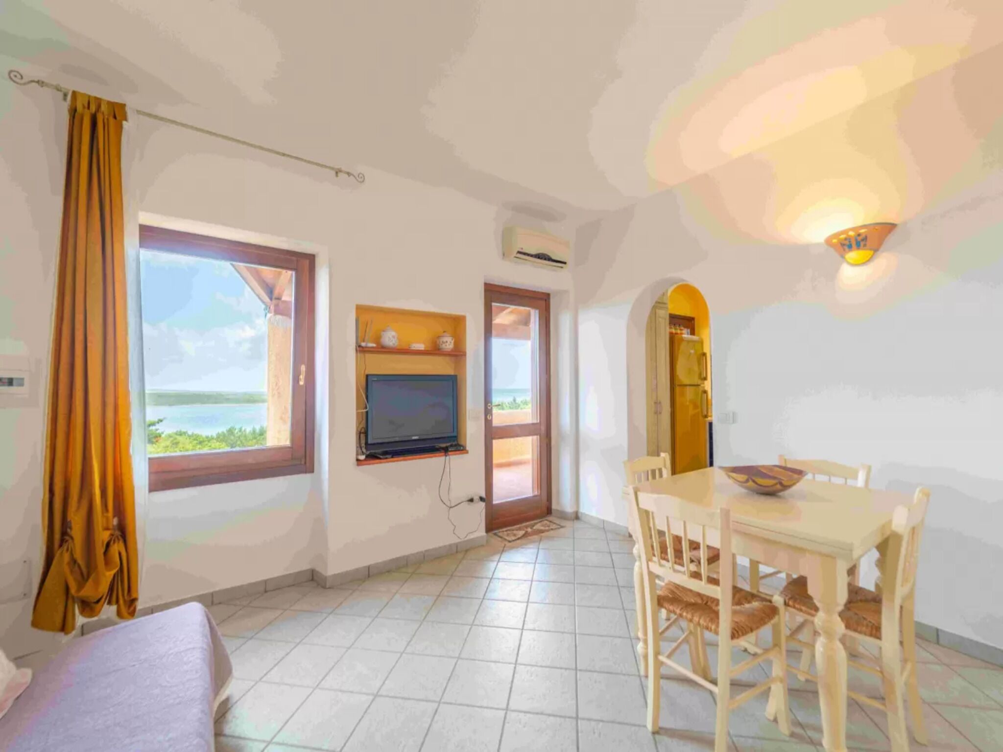A room with a bed, a TV, a dining table with chairs, a window with curtains, and a door leading to another room.
