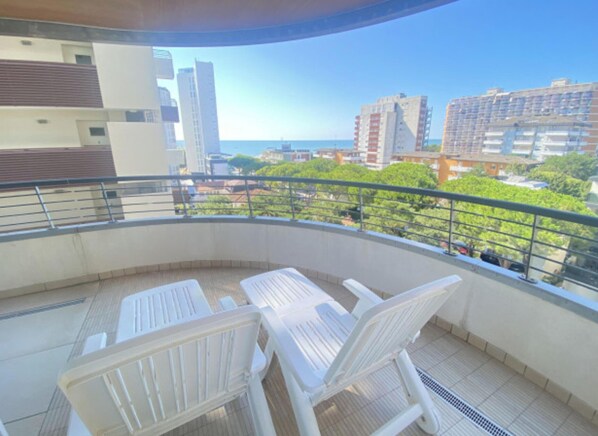 A balcony with two white chairs and a table, overlooking a cityscape with buildings and a clear sky. - Modern flat with panoramic view (Lignano Sabbiadoro)