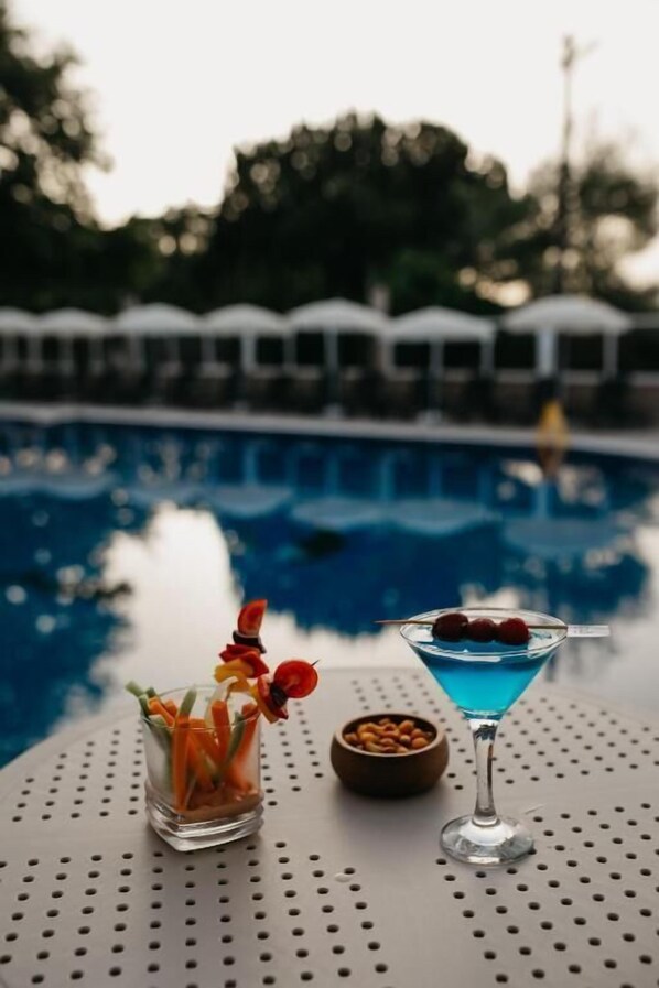 Une table au bord de la piscine avec un verre à martini et un verre de cocktail bleu, un bol de noix et un verre décoré d’une garniture de légumes.