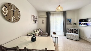 A dining area with a large wall clock, a table with chairs, a vase of flowers, and a sofa with patterned cushions. - STAY Heaven Apartment (Ayia Napa)