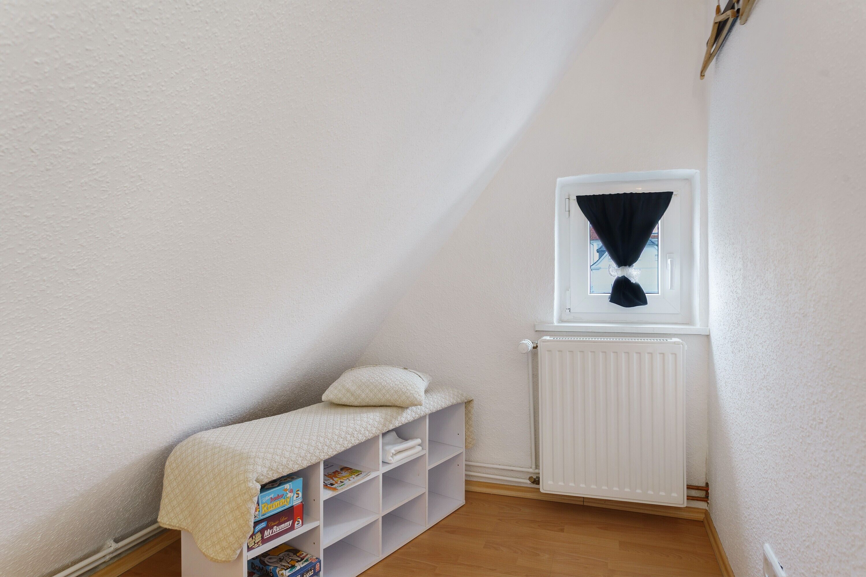A small, white-walled room with a sloped ceiling, a window with a black curtain, a radiator, and a shelf with books and a pillow.