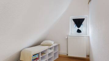 A small, white-walled room with a sloped ceiling, a window with a black curtain, a radiator, and a shelf with books and a pillow.