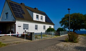 Une maison blanche dotée de panneaux solaires, une allée bordée d’une clôture et une vue dégagée sur la mer.
