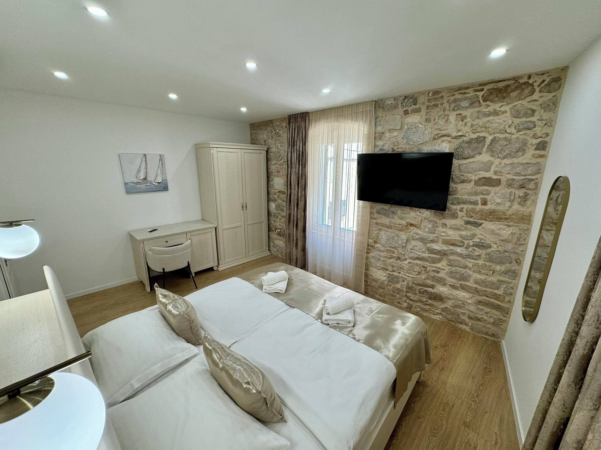 A hotel room with a bed, desk, chair, TV, and stone wall.