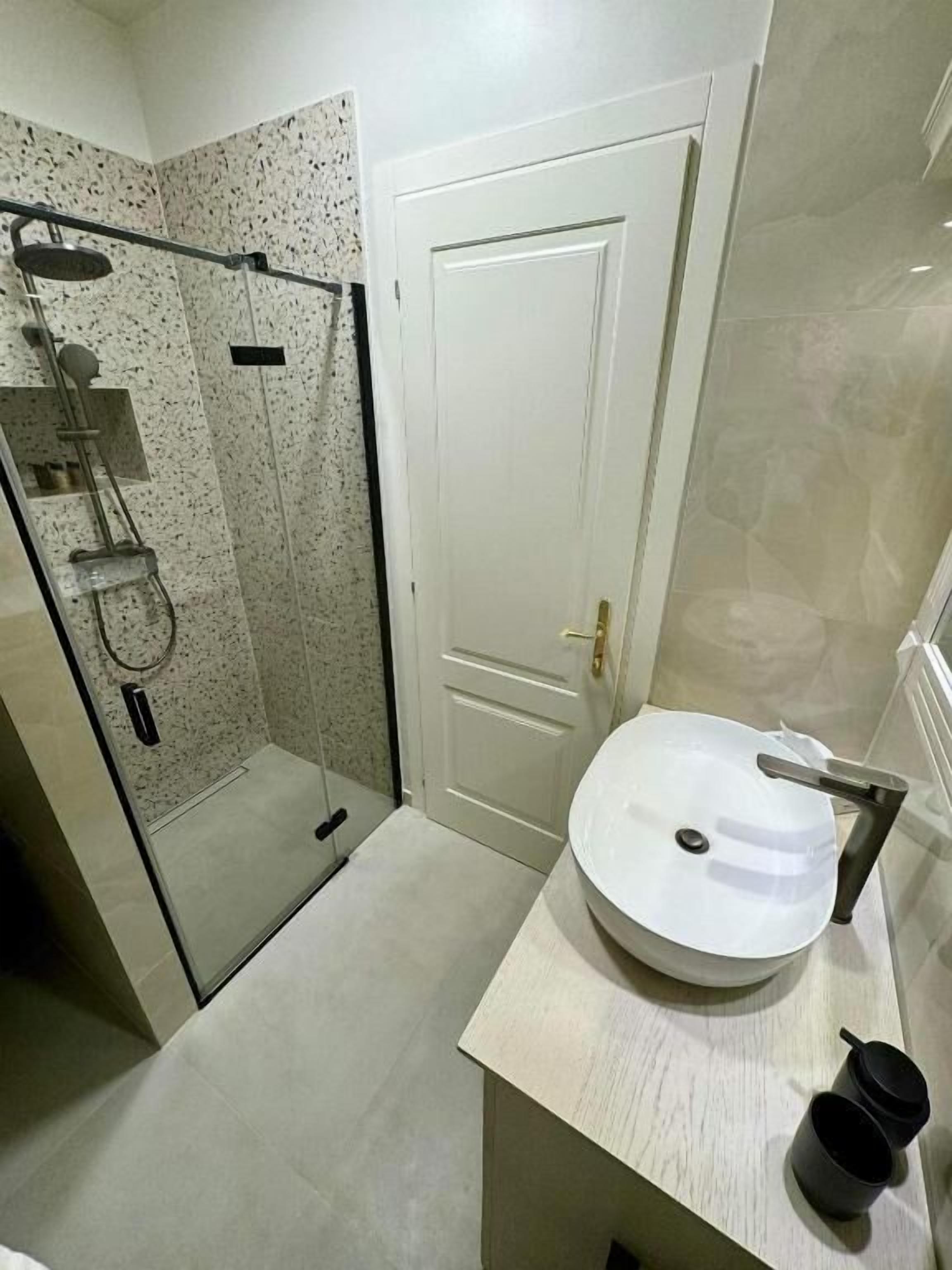 A bathroom with a glass-enclosed shower, a white door, and a round sink with a faucet.