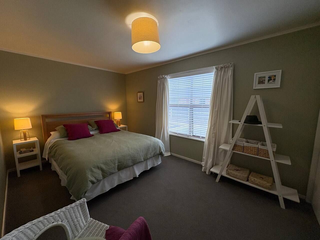 A bedroom with a bed, bedside lamps, a window with curtains, a white ladder shelf, and a small chair.