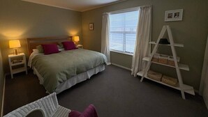 Een slaapkamer met een bed, bedlampen, een raam met gordijnen, een witte laddenplank en een kleine stoel.