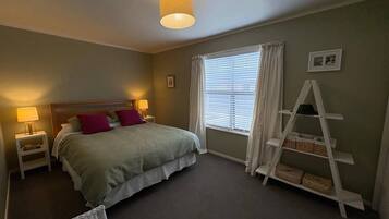 A bedroom with a bed, bedside lamps, a window with curtains, a white ladder shelf, and a small chair.