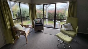 Een kamer met grote ramen, een rieten stoel, een comfortabele fauteuil en een kleine tafel.