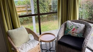 Een kamer met twee rieten stoelen, waarvan één met kussen en de andere met sierkussen, een klein rond tafeltje en een groot raam met gordijnen.
