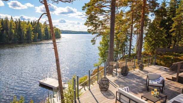 Ett trädäck med utemöbler, en hängstol och utsikt över en sjö som omges av träd.