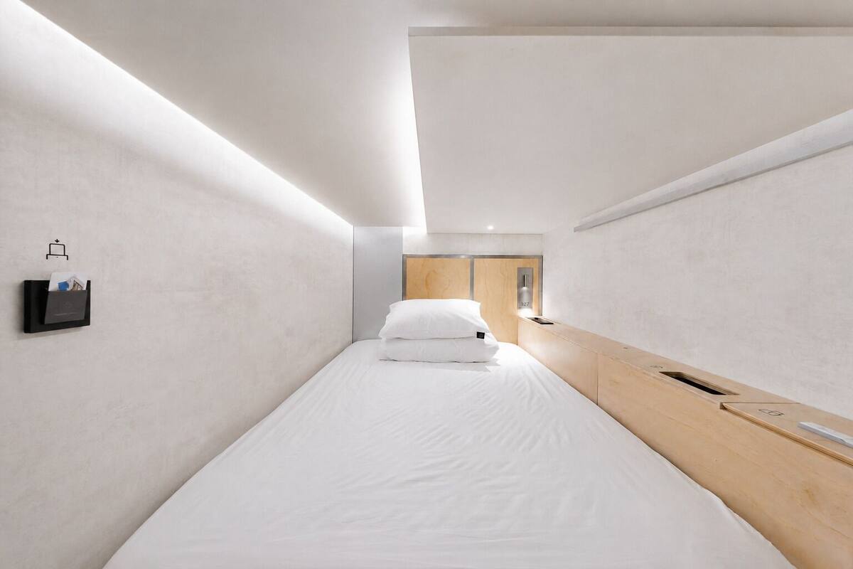 A modern hotel room with a single bed, a wooden headboard, a wall-mounted charging station, and a small bedside table.