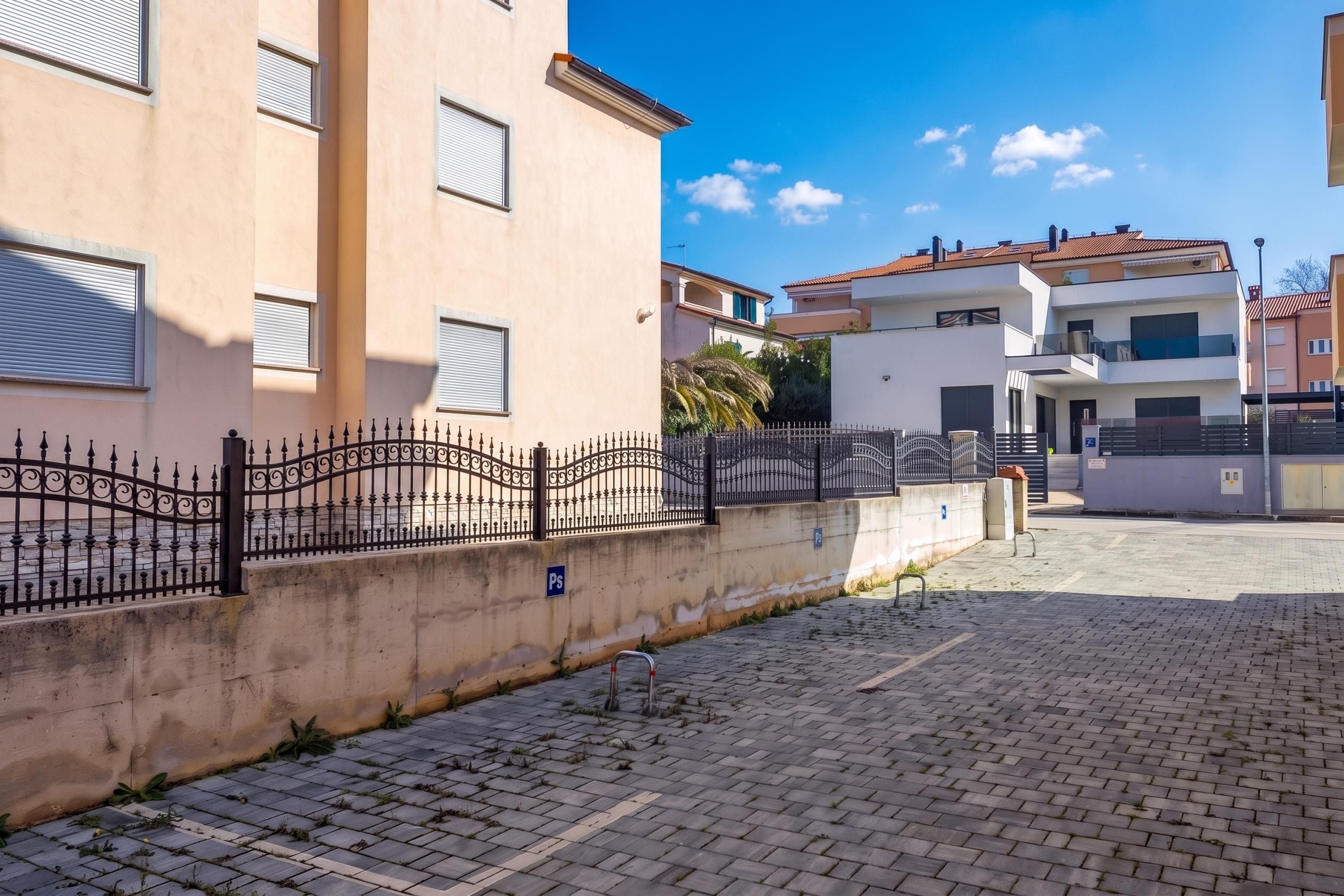 A paved courtyard with a metal fence, a parking sign, and buildings with balconies and windows.