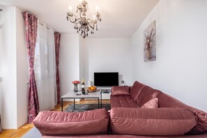 Un soggiorno moderno con un divano componibile, una TV a schermo piatto, un tavolo con un vaso e bicchieri da vino, e un lampadario.