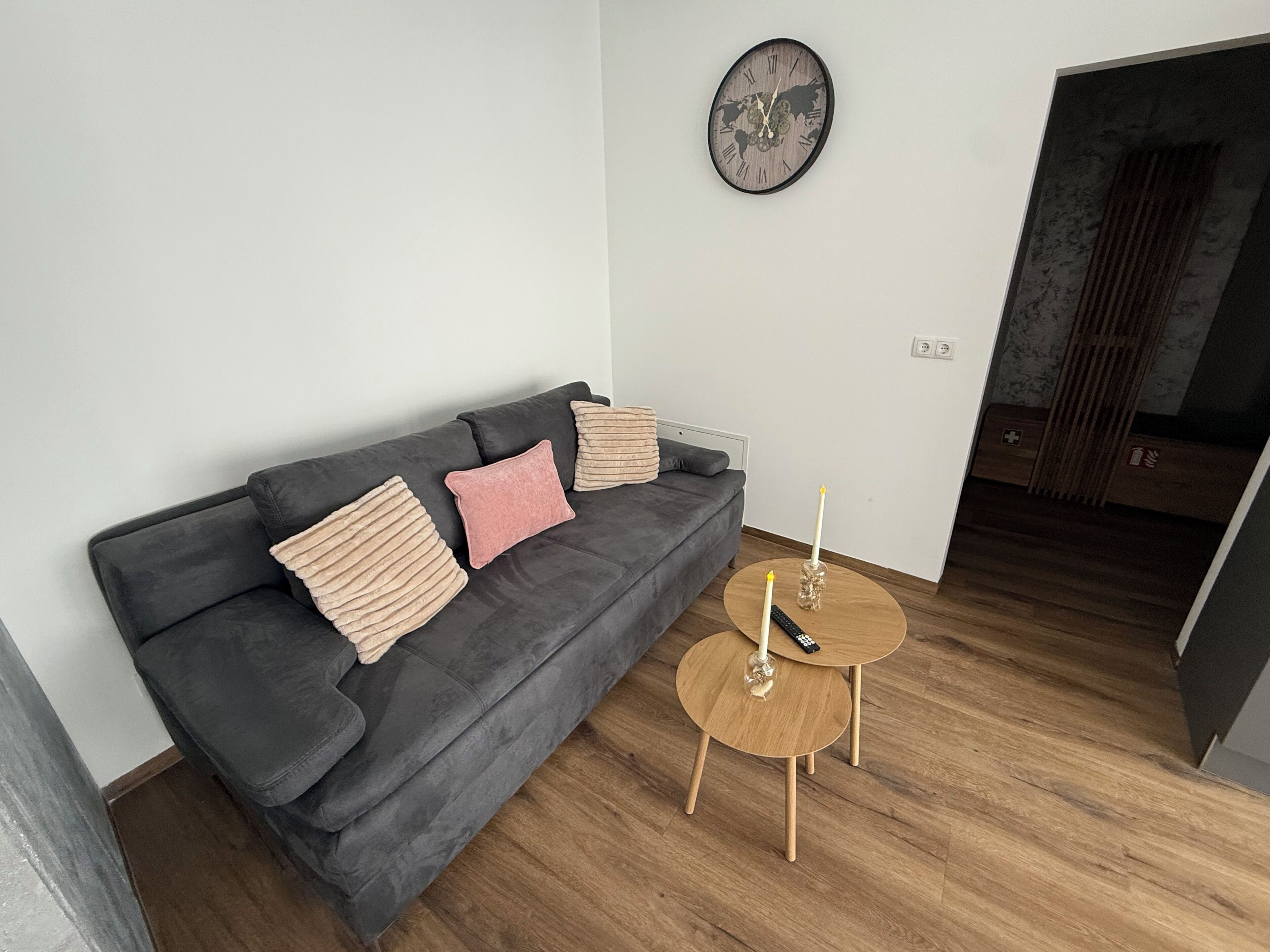 A modern living room with a dark grey sofa, two round wooden side tables, and a wall clock.