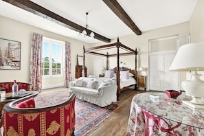 A bedroom with a four-poster bed, a sitting area with a red upholstered chair, a round glass table, and a large window with curtains.