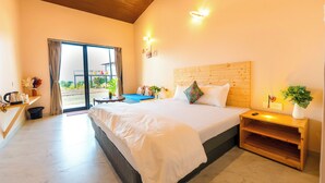 Habitación de hotel con una cama grande, un cabecero de madera, una mesita de noche con una planta, un escritorio con una silla y una puerta corrediza de vidrio al exterior.