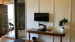 Ein Zimmer mit an der Wand montiertem Fernseher, einer Kaffeemaschine auf einem Holztisch, einem Mülleimer und einer Topfpflanze.