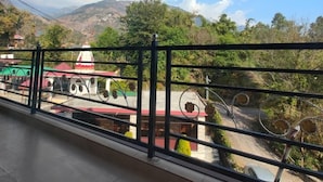 A balcony with a view of a mountainous landscape, a small structure with a red roof, and a metal fence. - AASRA PARADISE HOTEL (Dharamshala)