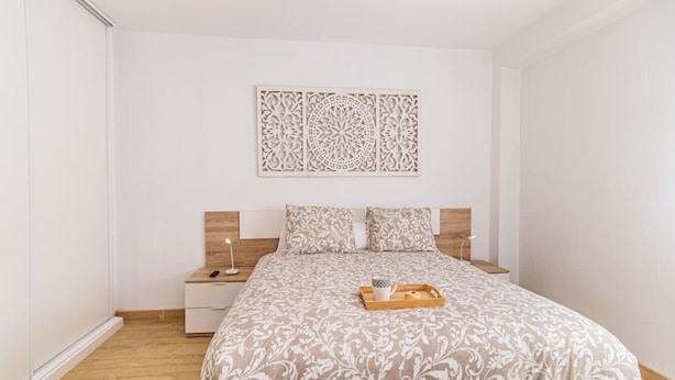 Un dormitorio con cama, mesitas de noche, una bandeja con objetos y un panel decorativo en la pared.