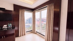 Quarto de hotel com painel de madeira escura, uma janela grande com cortinas, televisão de tela plana e frigobar.