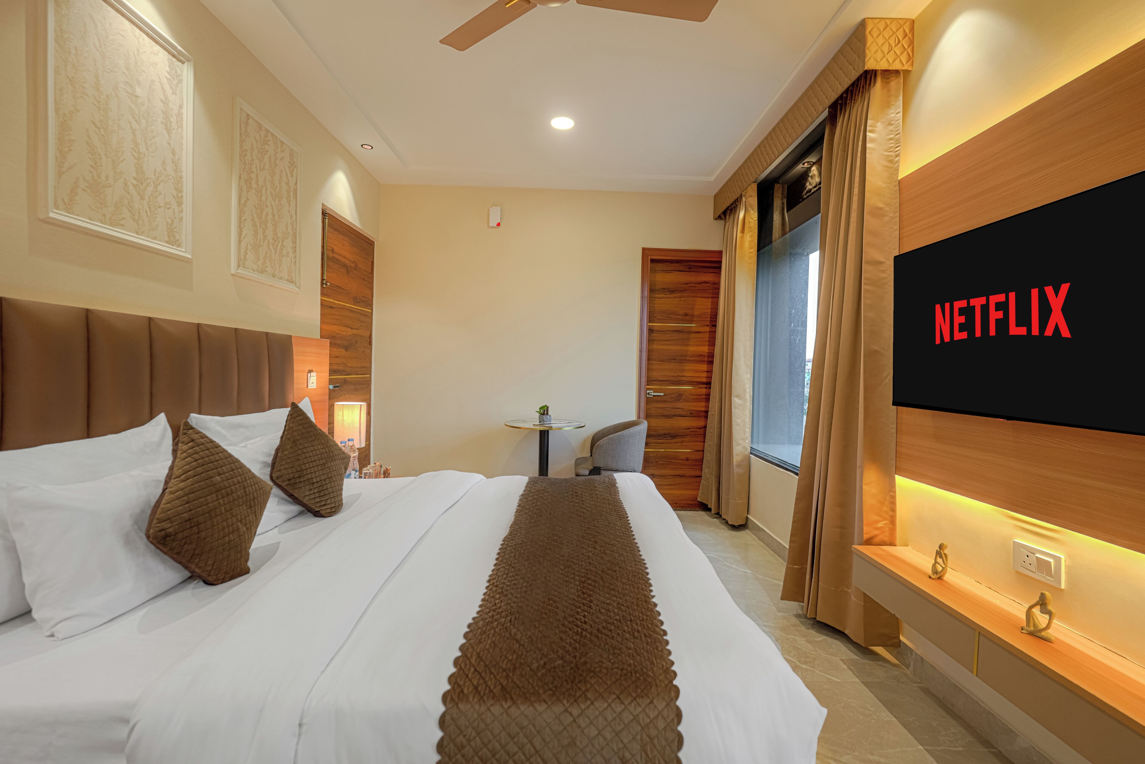 A hotel room with a bed, a TV mounted on the wall, a ceiling fan, and a small table with a chair.