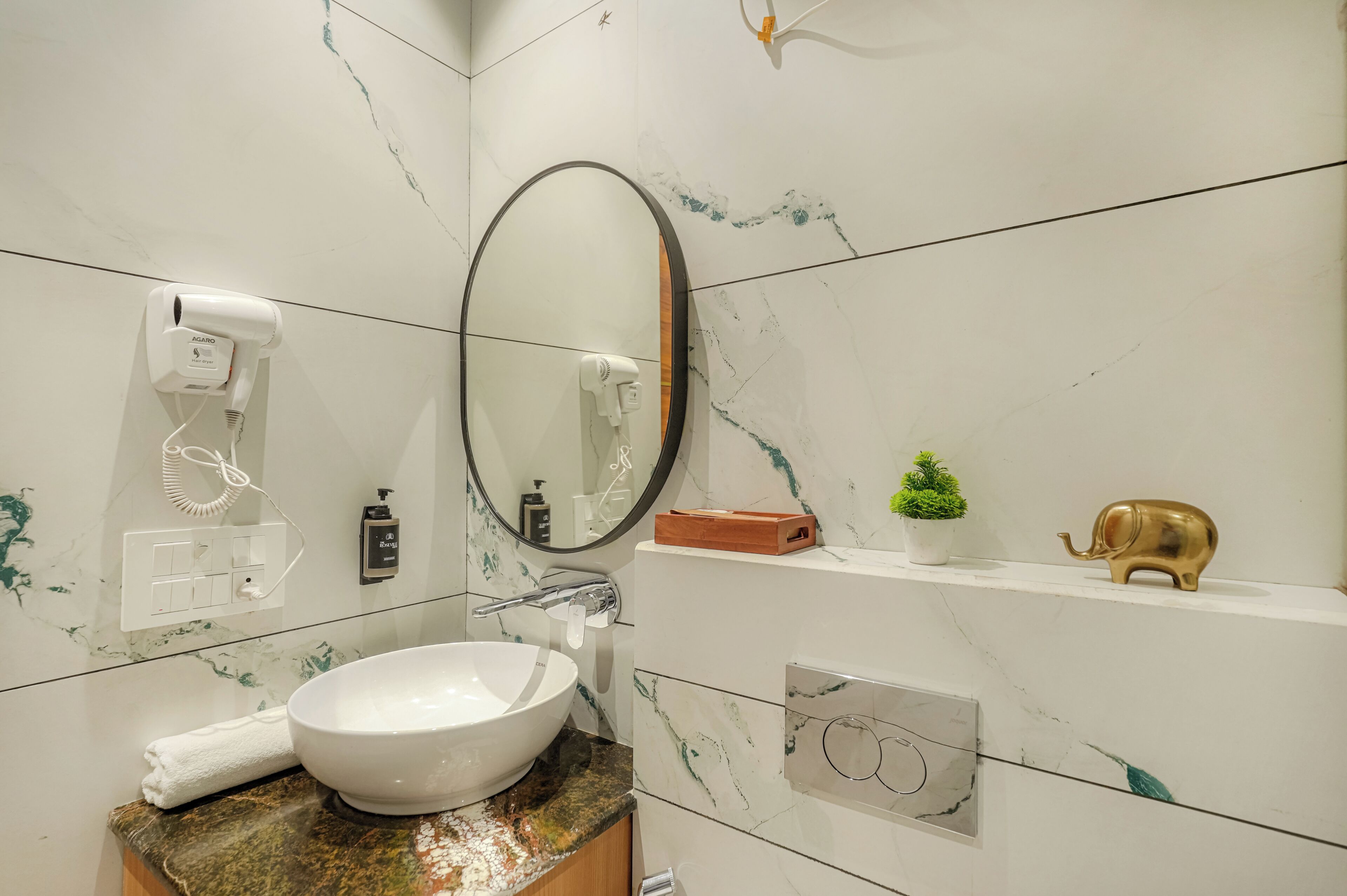 Een badkamer met een wit marmeren wastafel, een ronde spiegel, een haardroger, een klein plantje en een decoratief olifantje.