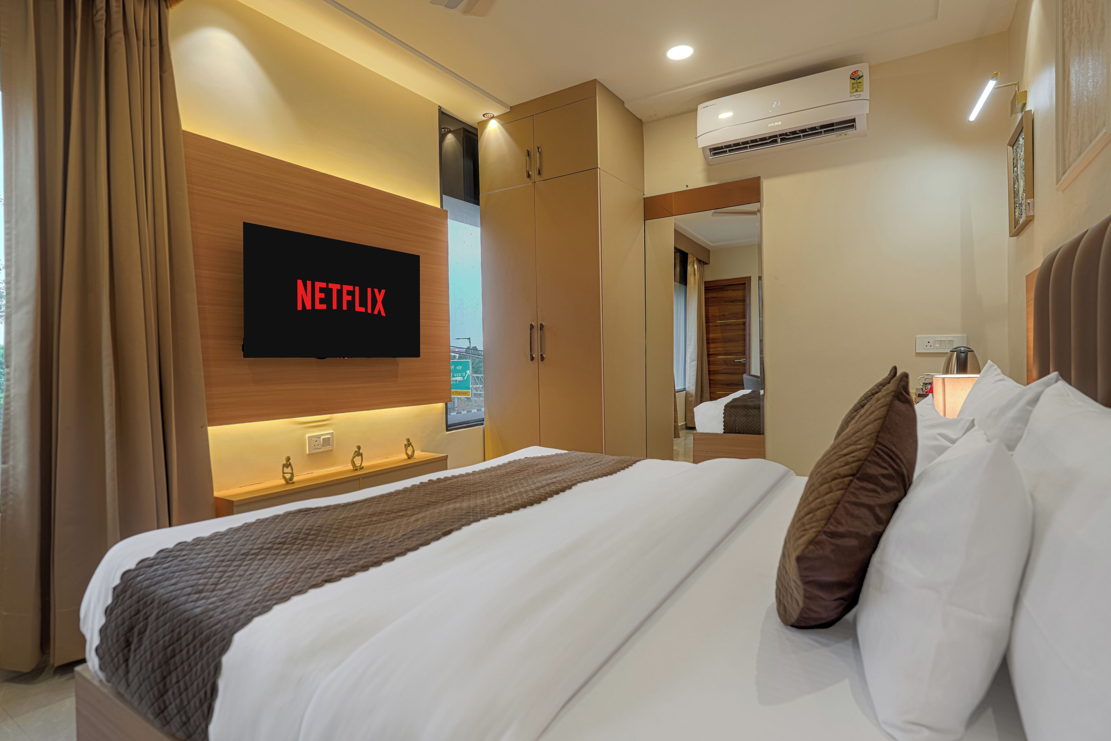 A hotel room with a bed, a TV mounted on the wall, an air conditioning unit, and a bedside table.
