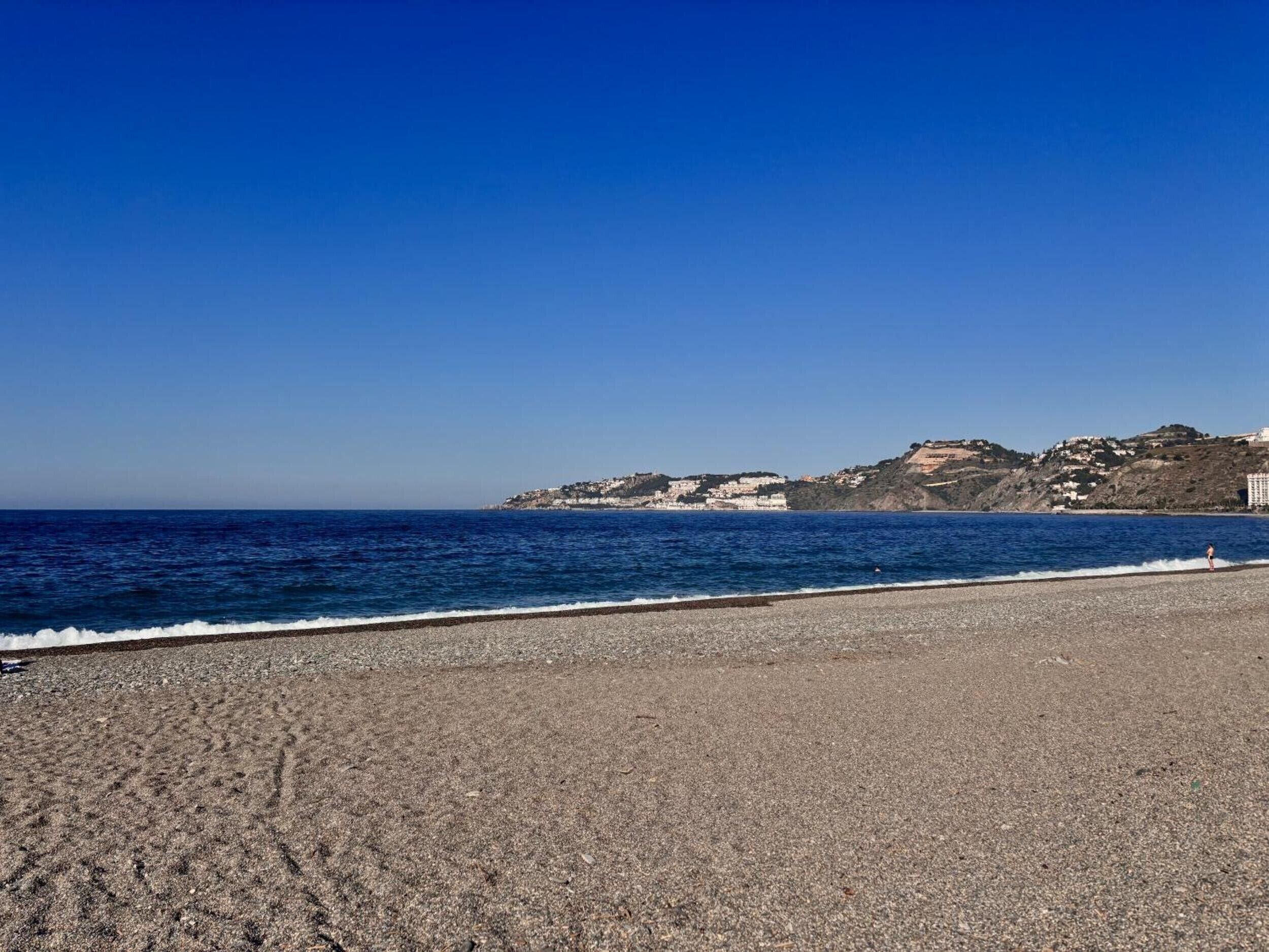 Una playa de canto rodado, cielo azul despejado y una colina distante con edificios.