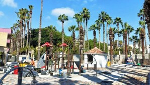 Ein Spielplatz mit Klettergerüsten, Palmen und einem kleinen Gebäude mit rotem Dach.