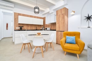 Eine moderne Küche mit weißen Schränken, einer Wand in Holzoptik, einem runden Esstisch mit weißen Stühlen und einem senffarbenen Sessel mit blauem Kissen.