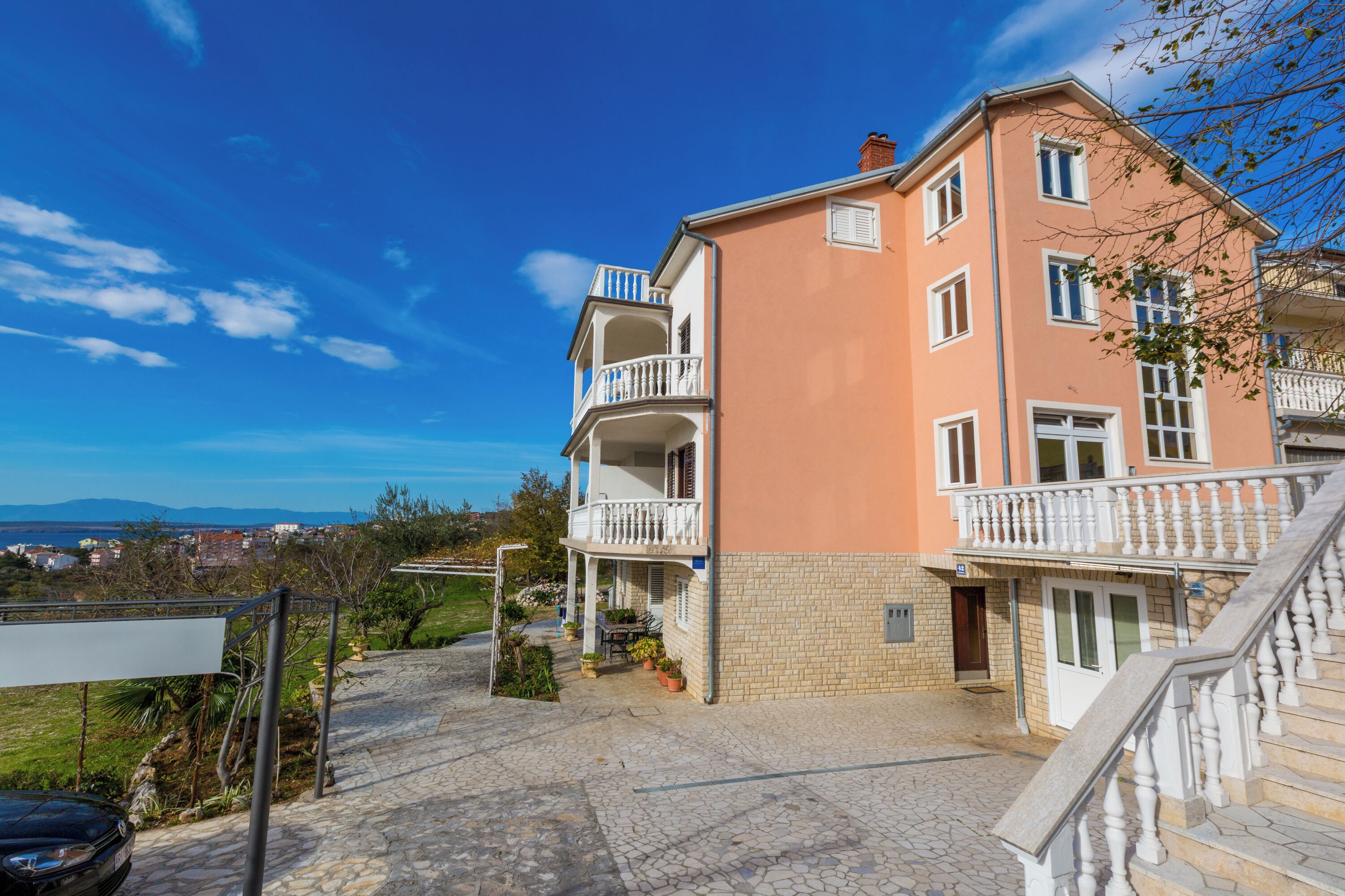 Un edificio de varios pisos con balcones, un acceso empedrado y vistas al paisaje.