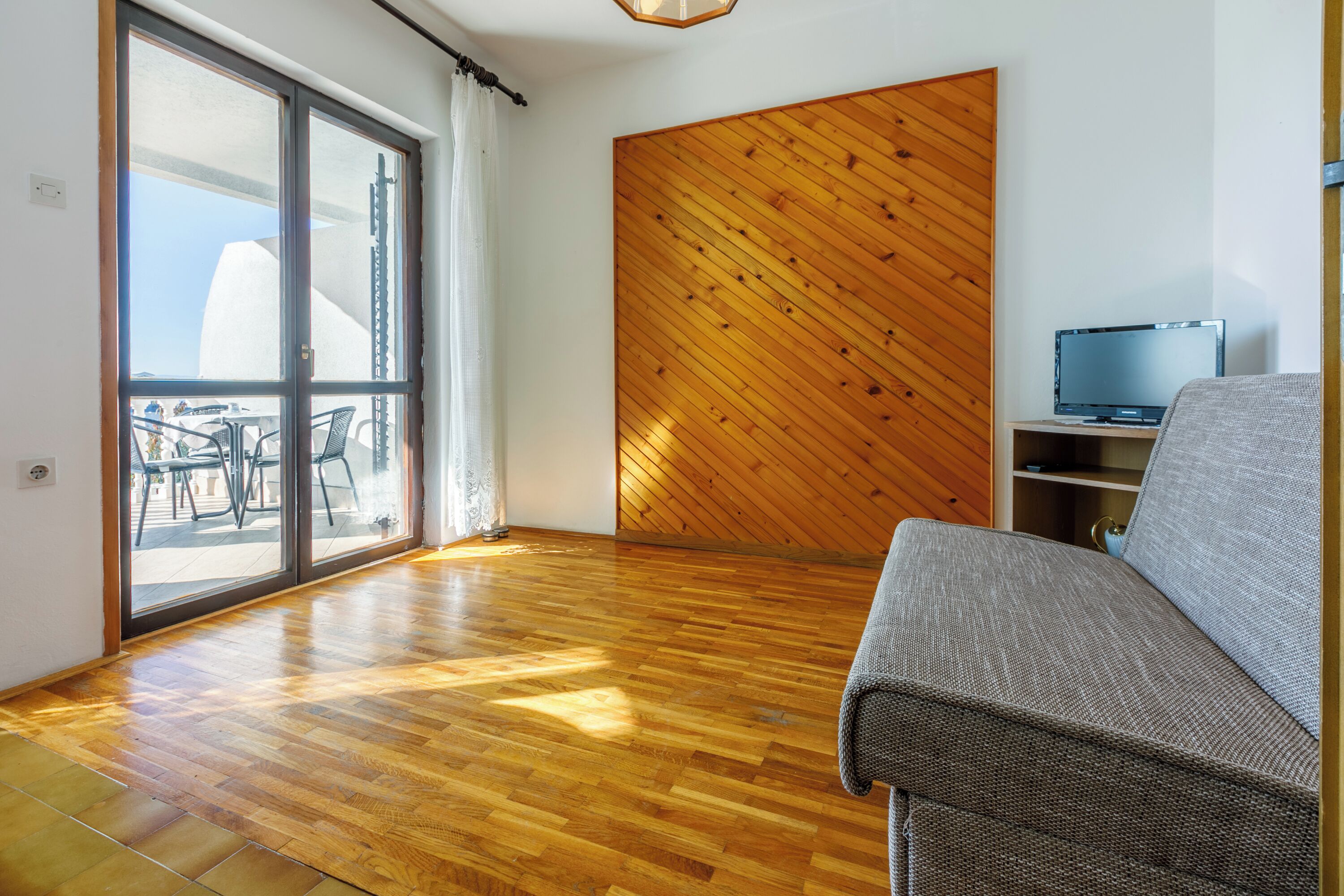 Habitació amb terra de fusta, un sofà, un televisor sobre un suport i una porta corredissa de vidre que dona a una zona exterior amb seients.