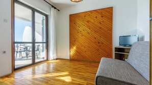 Une pièce avec un plancher en bois, un canapé, un téléviseur posé sur un meuble et une porte-fenêtre coulissante donnant sur un espace extérieur aménagé avec des chaises.