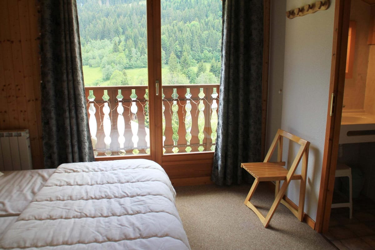 Une chambre à coucher avec un lit, une chaise en bois, une fenêtre donnant sur une forêt et une balustrade en bois.