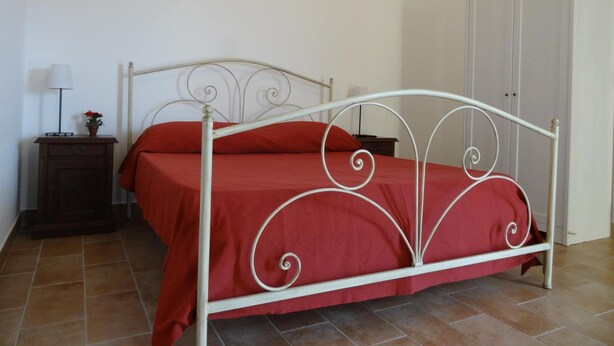 Un lit simple avec un cadre en métal blanc et une literie rouge, une table de chevet avec une lampe, et une armoire dans une pièce dont le sol est recouvert de carreaux.