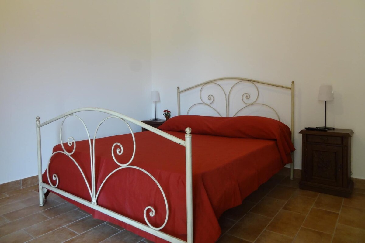 Ein Einzelbett mit Metallgestell und roter Bettwäsche, ein kleiner Nachttisch mit Lampe sowie ein Schrank an der Wand.