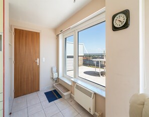 Ein Zimmer mit Holztür, Fenster mit Blick auf einen Balkon, Heizkörper und einer Wanduhr.