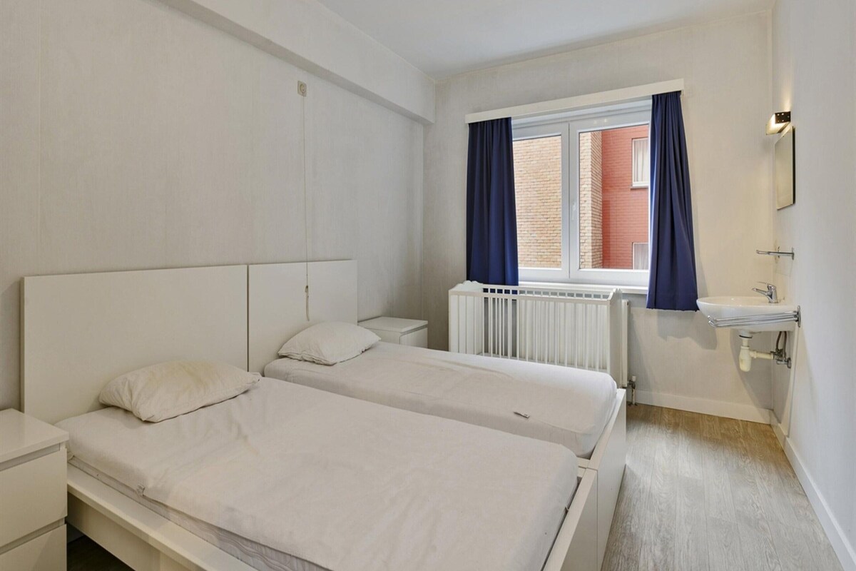 Ein Schlafzimmer mit zwei Betten, einem Fenster mit Vorhängen, einem Heizkörper, einem Waschbecken und einem Spiegel.
