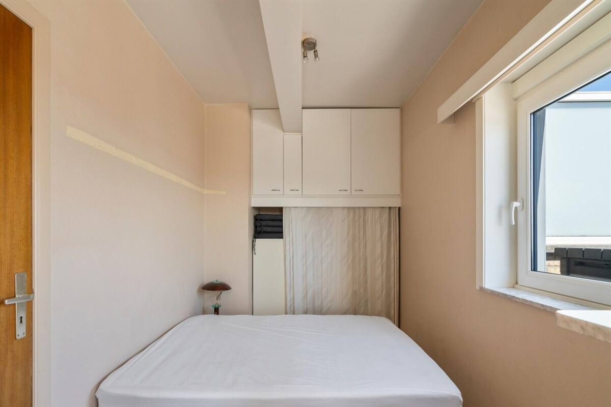 Ein kleiner, schmaler Raum mit einem Einzelbett, einem Fenster und einer Tür.