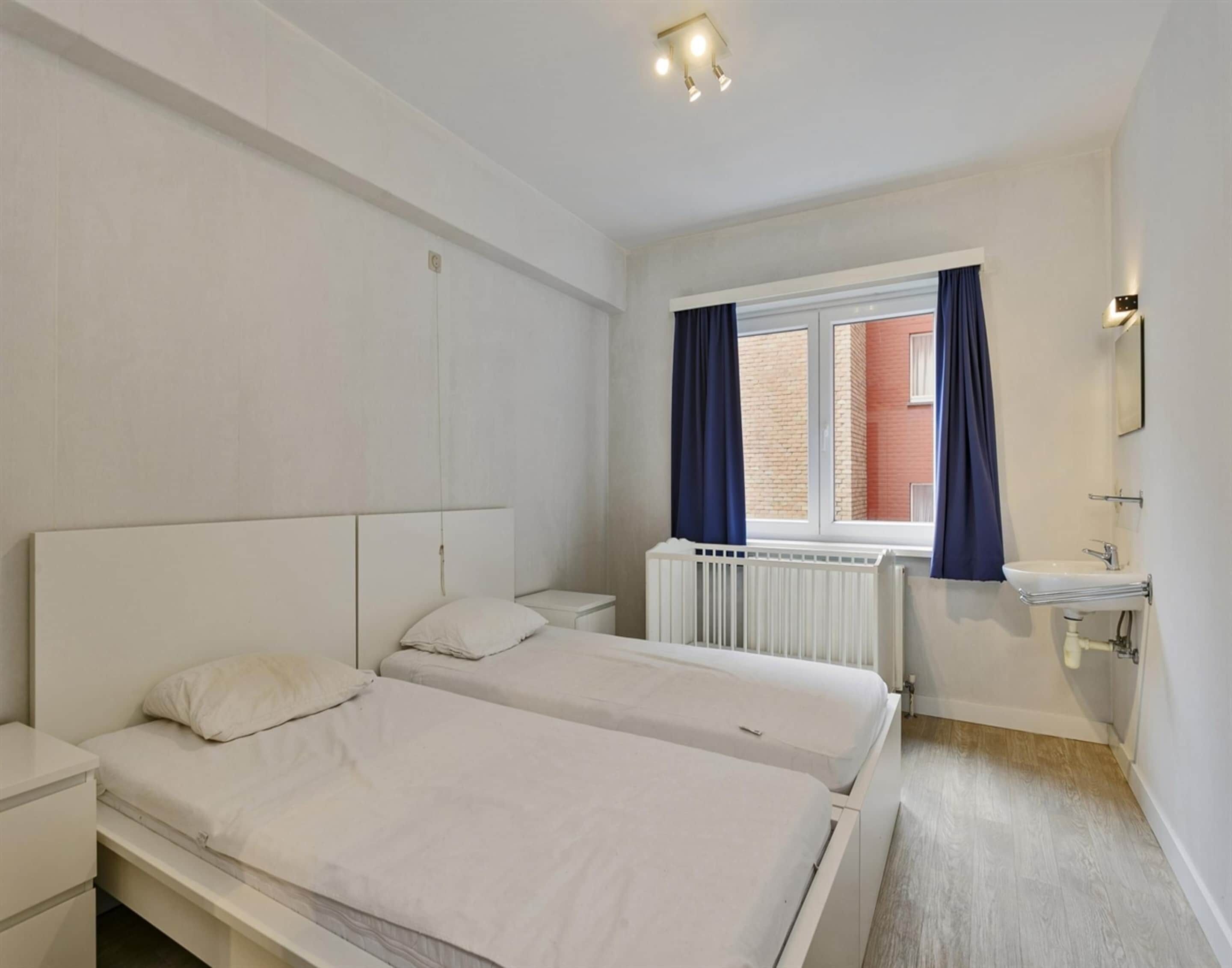 A bedroom with two beds, a window with curtains, a radiator, a sink, and a mirror.