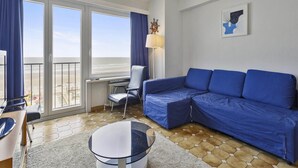Ein modernes Wohnzimmer mit einem blauen Sofa, einem Glastisch, einem Schreibtisch und einem Sessel. Große Fenster mit Vorhängen, eine Deckenleuchte und ein gerahmtes Bild sind an den Wänden zu sehen.