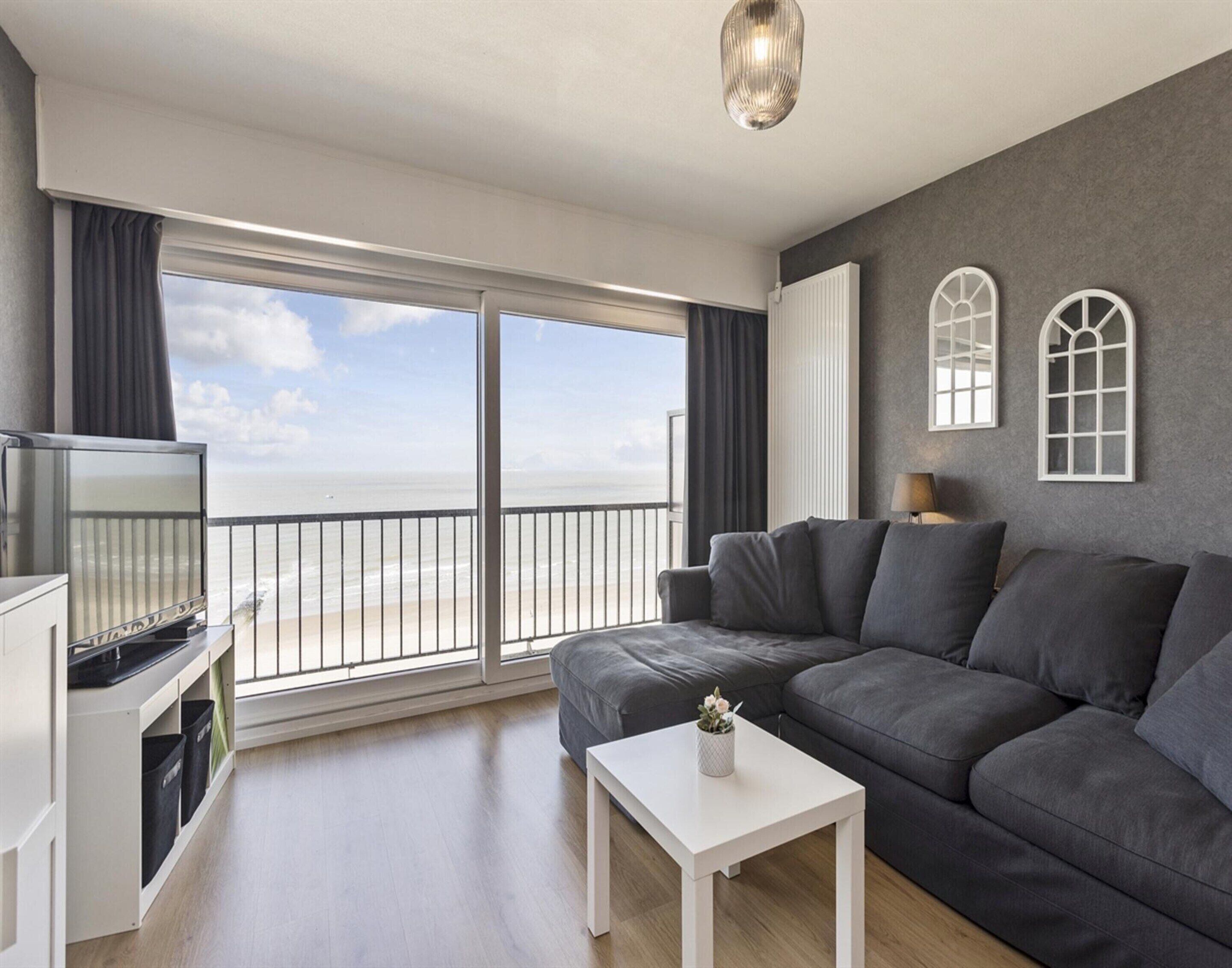 A modern living room with a grey sofa, a white coffee table, and a TV on a white cabinet. There are large windows with a view of the beach.