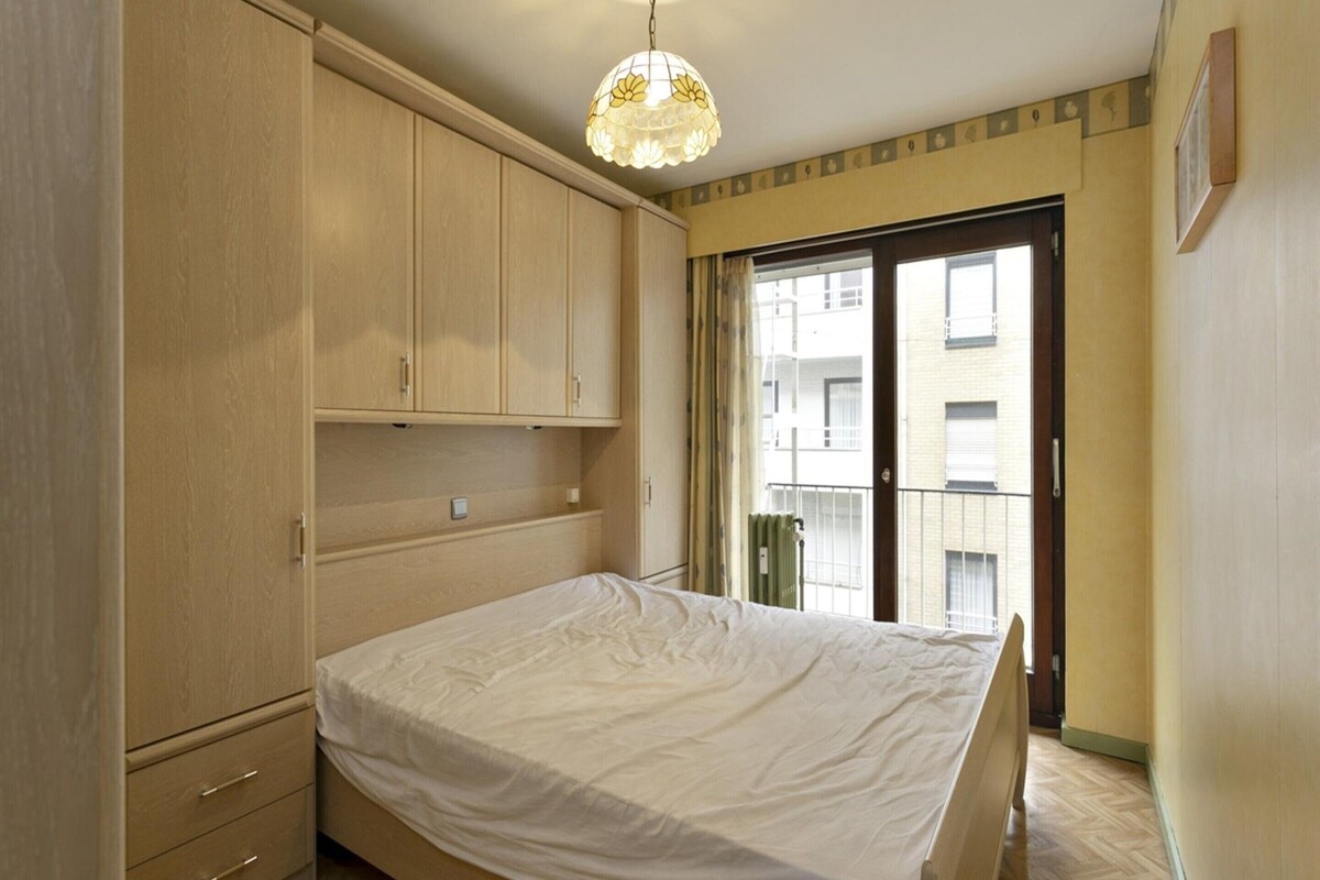 Ein Schlafzimmer mit Bett, Holzschrank und Blick auf ein Gebäude durch eine Glastür.