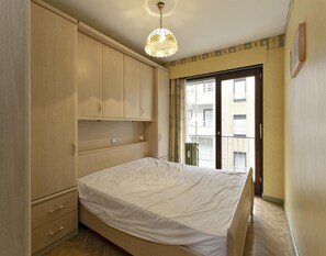 A bedroom with a bed, wooden wardrobe, and a view of a building through a glass door. - Apartment in Blankenberge Near the Pier (Blankenberge)
