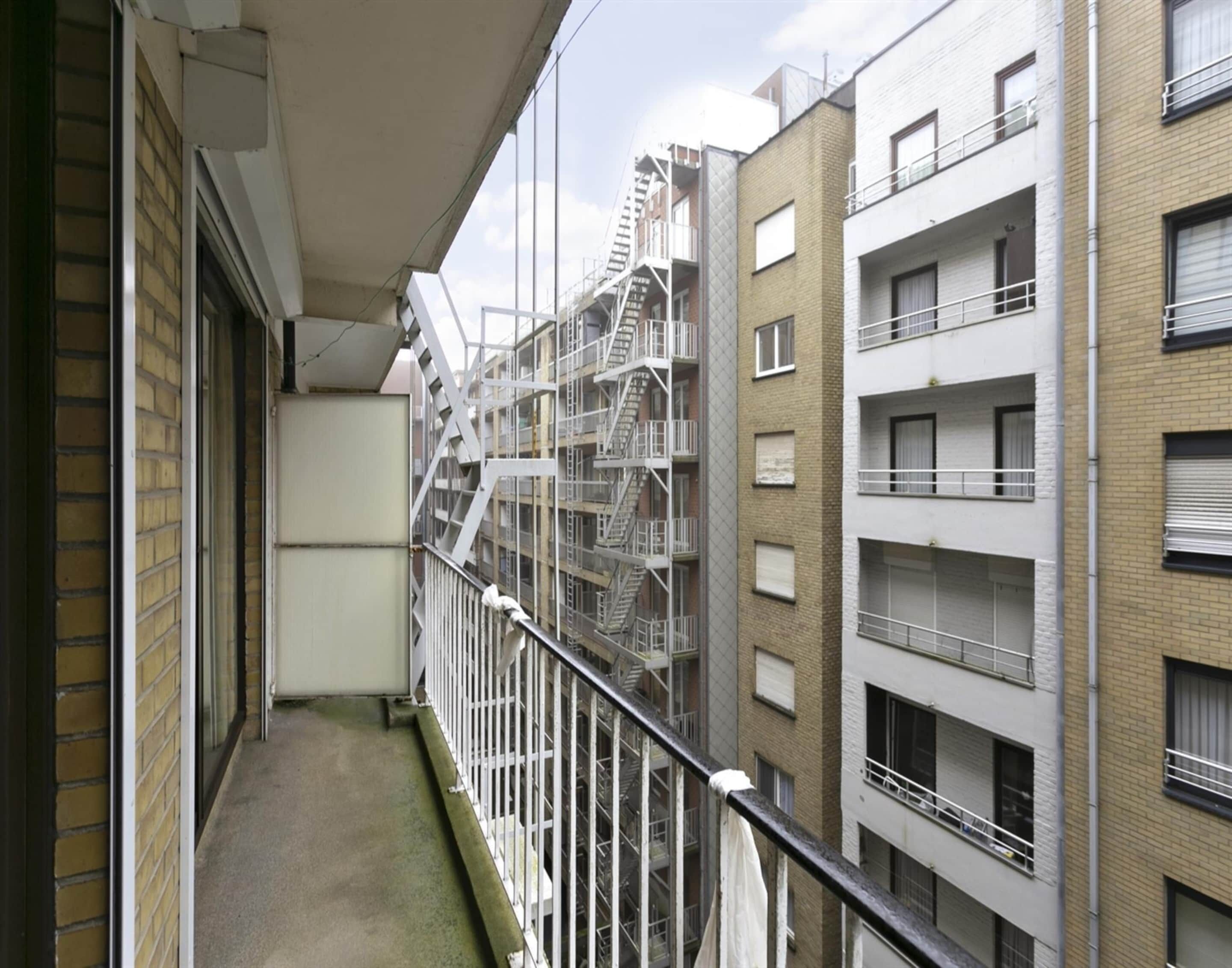 A narrow balcony with a metal railing, adjacent to a multi-story building with visible fire escapes.