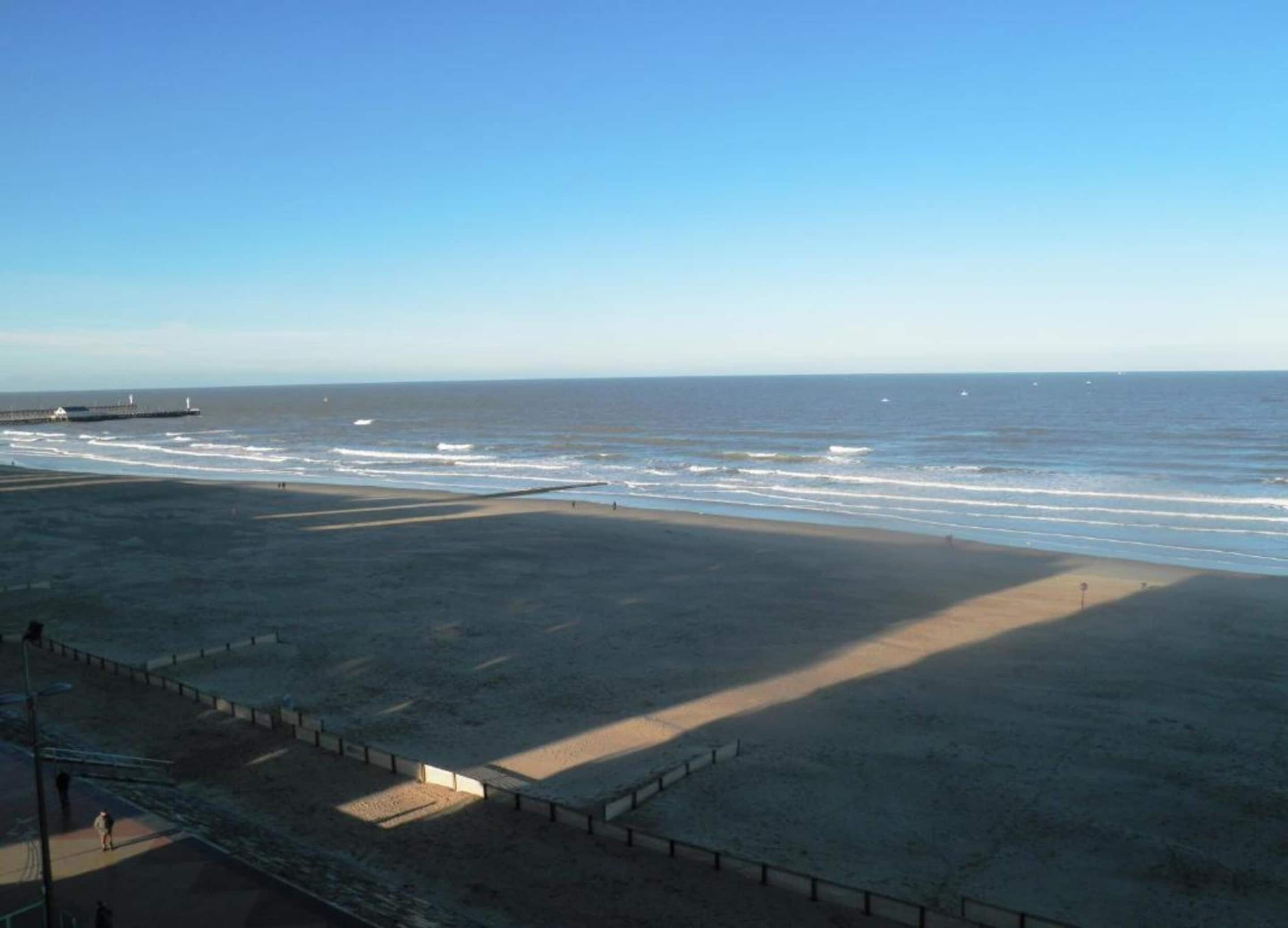 A beach with a pier in the distance, a fenced area, and a clear sky.