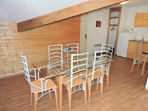 Uma sala de jantar com mesa de vidro e cadeiras brancas, piso de madeira e cozinha equipada com geladeira e máquina de café.